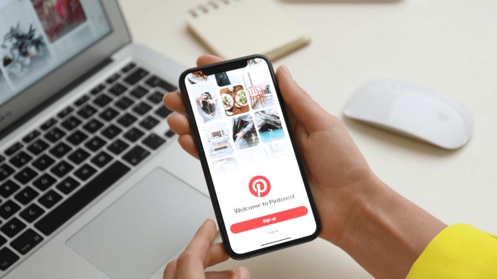 Woman holding a mobile phone in front of her laptop with the Pinterest app on open on her phone. 