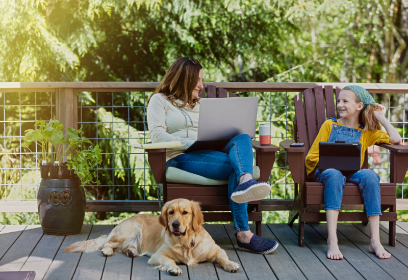 A mompreneur working on her laptop outside on a nice day on a deck with her daughter and golden retriever.