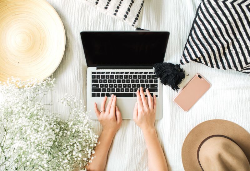 Mompreneur typing on a laptop with an aesthetic background of flowers, pillows, and hats.